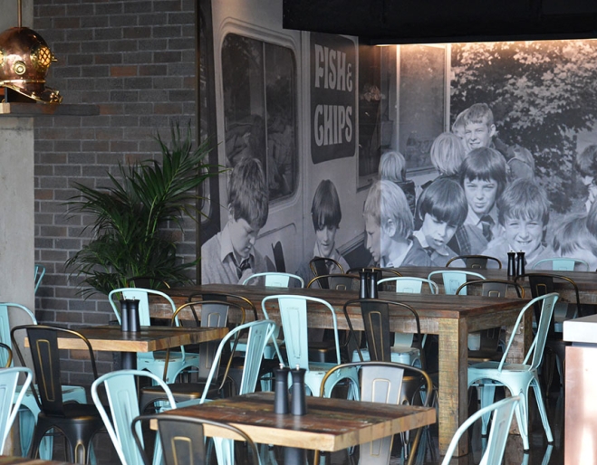 Wooden tables and blue chairs against a black and white mural wall inside Sea Fuel Fish & Chips, West End