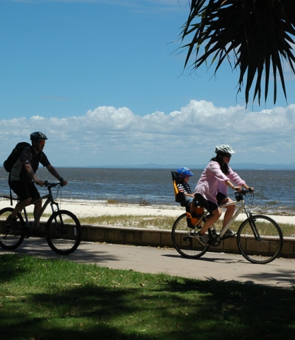 Family cycling on cycle path on Bribie Island with Moreton Bay ocean in background.
