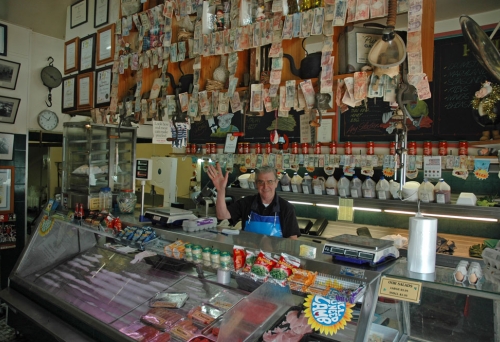 Butcher waving behind counter covred in foreign bank notes inside Annerley Meats in Brisbane.