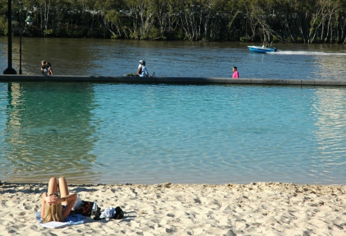 Woman lying on Streets Beach looking at speed boad on Brisbane River and mangroves.