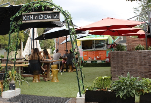 Diners at alfresco café on Astroturf with a mix of tables and chairs and a retro caravan kitchen at Kith 'n' Chow Mt Gravatt