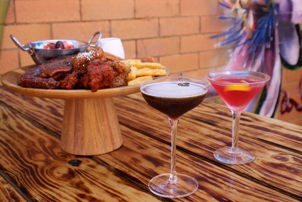 Two cocktails and a wooden platter of meat, fries and fruit salad on a wooden table at King of the Wings, Stafford